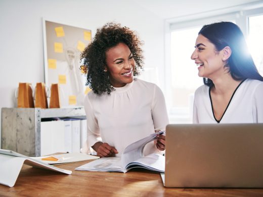 Two happy attractive young multiracial businesswomen laughing together as they sit working in a bright high key office on paperwork and a laptop computer